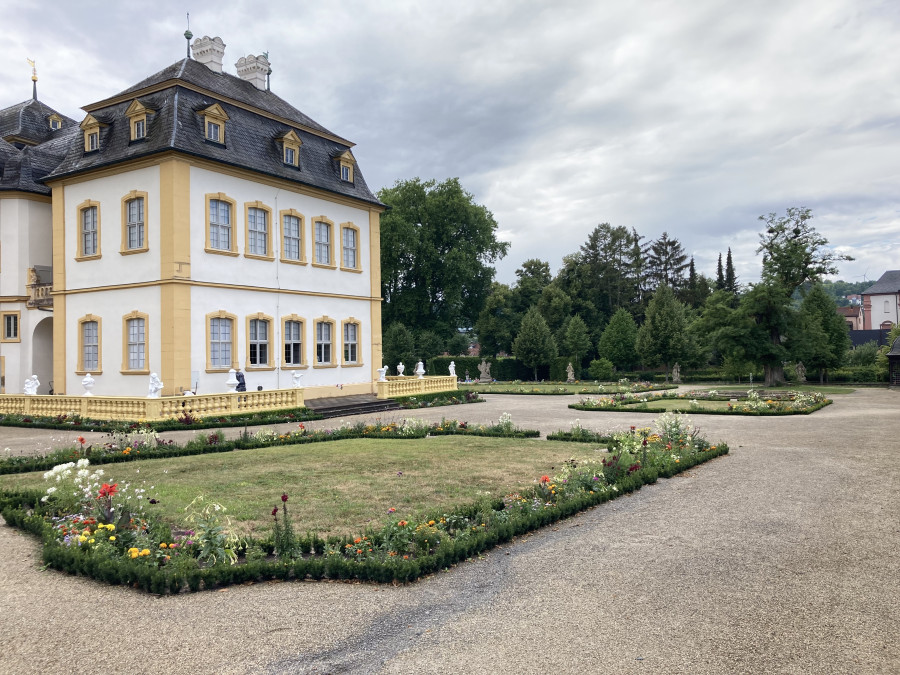 Das Bild zeigt einen Ausschnitt des Veitshöchheimer Hofgartens, links ist ein Teil des Schlosses zu sehen. Im Vordergrund Kieswege mit Grasinseln, die von buntblühenden Pflanzen gesäumt werden. Die Grasinseln wurden mit trockenheitsverträglicher Rasenmischung bearbeitet und zeigen teilweise braune vertrocknete Stellen