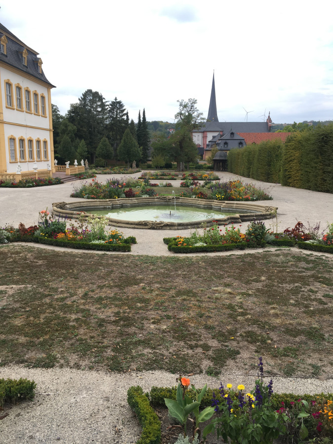 Das Bild wurde im Hofgarten Veitshöchheim im September 2020 aufgenommen. Im Vordergrund sind durch Trockenheit großflächig geschädigte braune Rasenflächen mit bunten Blumen und Fontänenbecken zu sehen, links ein Teil des Schlosses