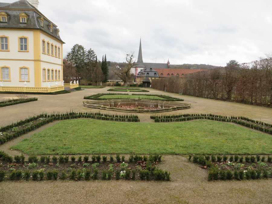 Links im Bild ist ein Teil des Veitshöchheimer Schlosses im Februar 2023 zu sehen, in der Mitte des Bildes grüne Rasenflächen im Parterre, die von jungen, noch kleinen Pflanzen umsäumt sind