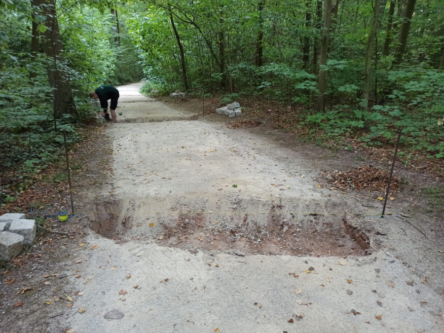 Ein von grünen Bäumen umstandener Parkweg mit einer über die ganze Breite des Weges verlaufenden Baugrube