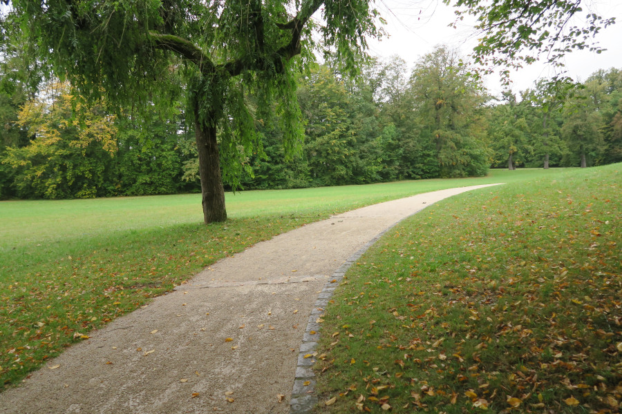 Baumbestandene grüne Wiesen mit gelbem Laub, die von einem hellgrauen Weg durchzogen werden. Dieser hat eine Querrinne aus Granitstein, damit das Wasser nach starkem Regenfall ablaufen kann