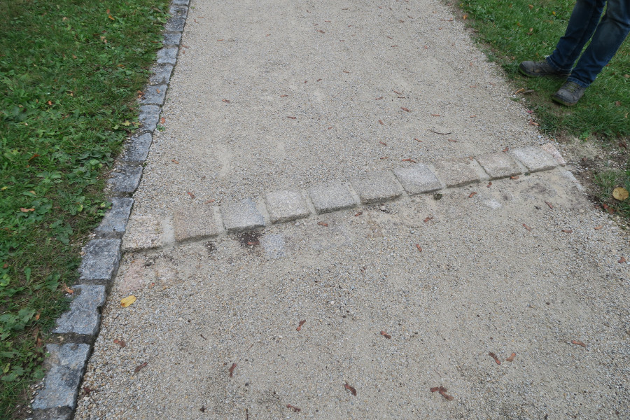 Hellgrauer Weg mit einer Querrinne aus Granitquadersteinen zu Entwässerung nach starkem Regenfall im Park Rosenau. Der Weg ist an einer Seite mit rechteckigen Steinen eingefasst, links und rechts des Weges ist Rasen.